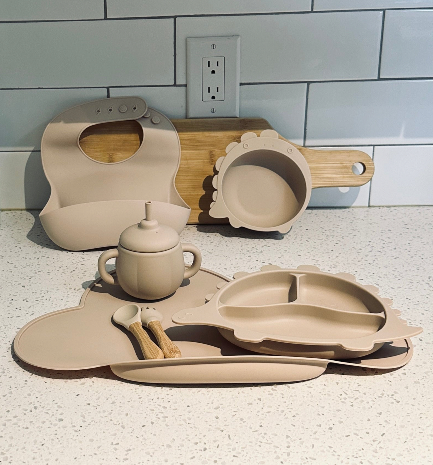 Neutral beige DinoFeast silicone feeding set for toddlers, including dinosaur plate, bowl, bib, utensils, and cup on a kitchen counter.