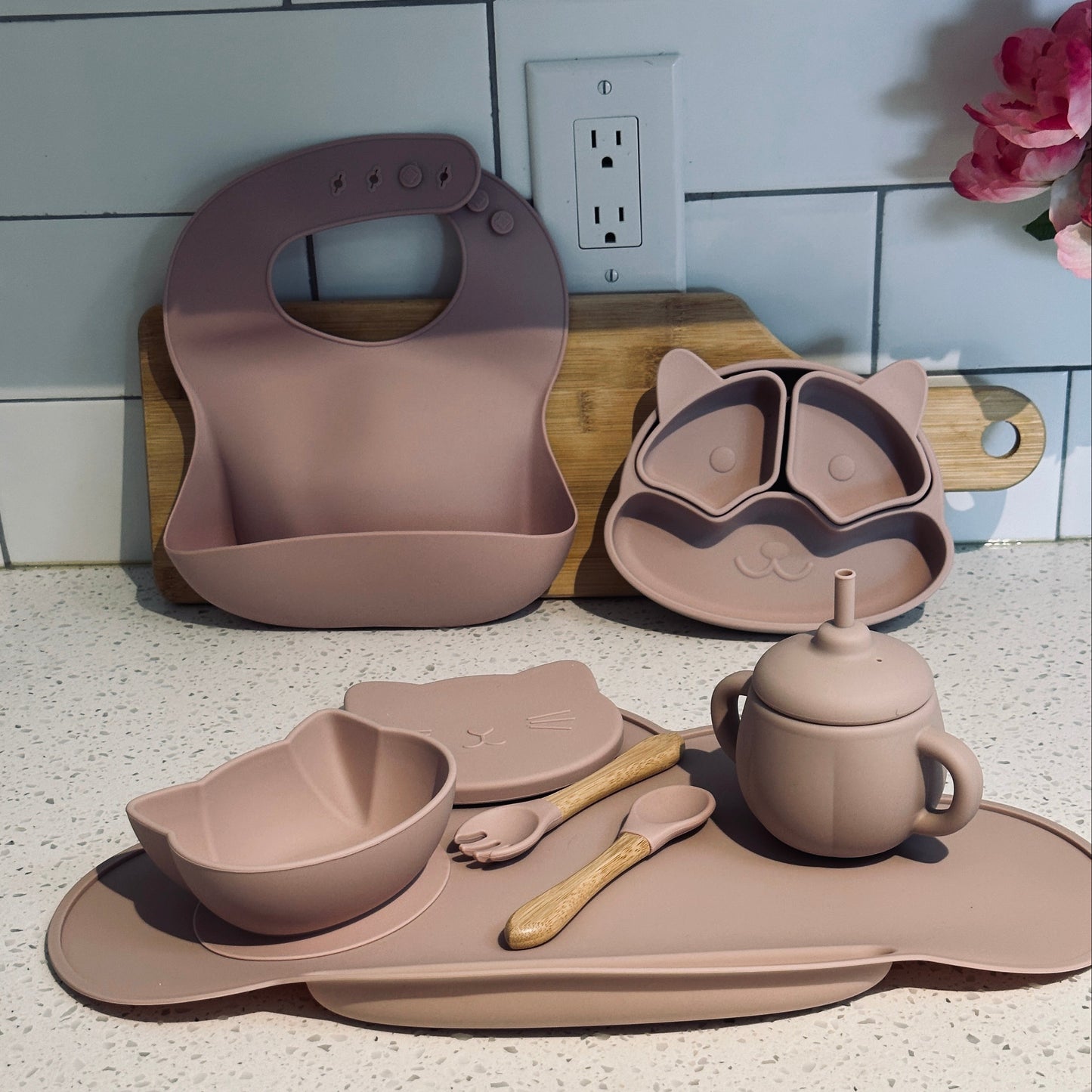 Dusty rose AnimalFeast silicone baby feeding set with raccoon plate, cat bowl, bib, utensils, and sippy cup on a kitchen counter.