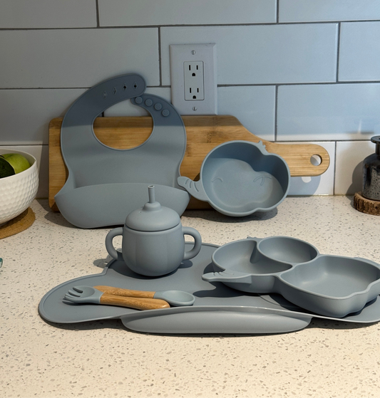 Stone blue DinoFeast silicone baby feeding set on kitchen counter, including dinosaur plate, bowl, bib, cup, and utensils.
